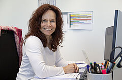Frau Läufer sitzt an ihrem Schreibtisch und lächelt in die Kamera. Im Hintergrund eine farblich abgesetzte Tabelle.